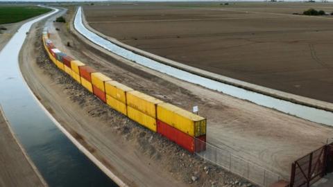 Photo of yellow and red shipping containers stacked in twos to fill a gap in an unfinished wall on the US-Mexico border
