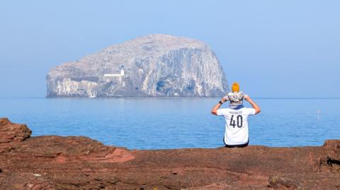 A man wearing a football shirt and with his child sitting on his shoulders looks across at an island on a clear, sunny day.