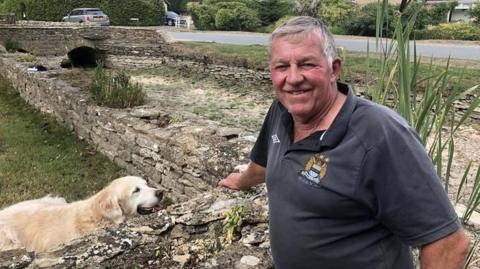 Martin Carter wearing a black polo shirt stood by the dried up River Thames in Ashton Keynes. A cream-coloured labrador dog is close by him, with a small stone wall separating them