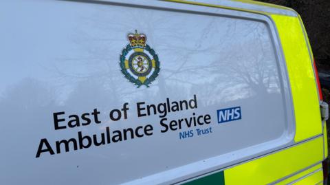 A close up shot of the side of a small ambulance. It is white and neon yellow, with these words on the panel: East of England Ambulance Service NHS Trust. Their logo, which is a Roman-style wreath, is above the words.