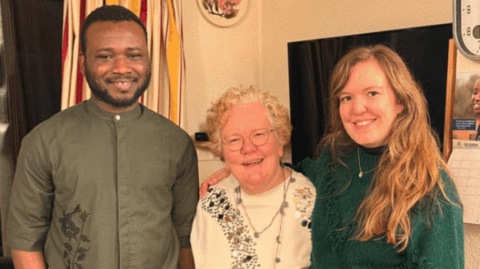 A young man, an older woman and a young woman stand together smiling with their arms around each other in a living room 