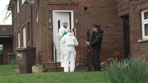 Policeman in black uniform and cap speaks to two forensic officers in white kit as they stand outside the entrance to the brick house. A garden lawn and plants are at the front with wheelie bin at the side of the house.