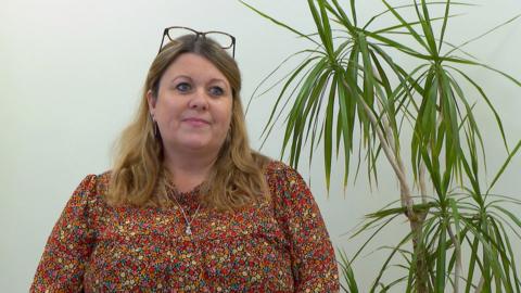 Nikki Laine, who is wearing a flowery blouse with red, orange and yellow flowers on it, sits next to a green house plant.