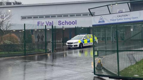 A photo taken from the school gates shows a police car parked in front of a school building. Purple letters on the white building read "Fir Vale School", although the E has fallen off.