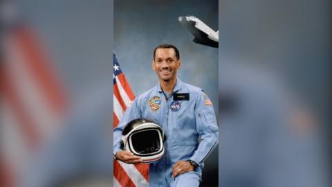 Charles Bolden poses in a blue Nasa uniform, holding a helmet. He stands in front of a US flag and an image of a space shuttle