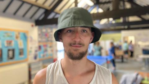 James, a 20-year-old wearing a green bucket hat and white vest, looks directly at the camera. He has a brown beard and moustache.