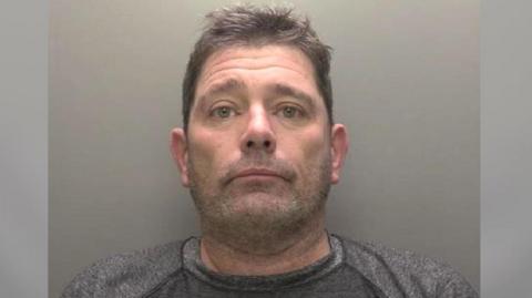 A police custody picture showing the head and shoulders of a man with short brown hair and stubble wearing a grey T-shirt against a grey backdrop. 
