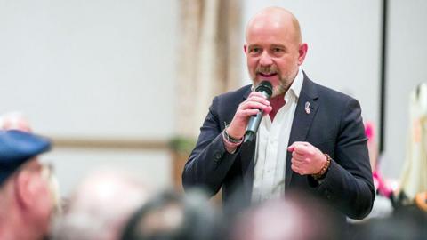 Steve Hilton speaks at a campaign event. He holds a microphone, and wears a black blazer with a pin in the shape of California.