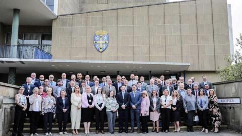 More than 50 people - men and women - lining the steps in front of Durham County Hall - part of the building can be seen behind them. It is of beige coloured stone with a plaque of the the county crest.