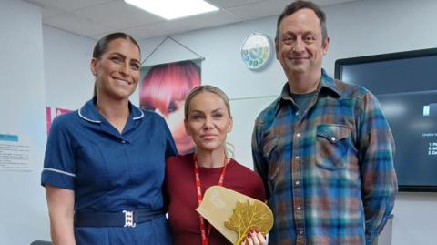 The image shows three people smiling and posing for a picture. The woman in the middle is blonde and wearing a red top and lanyard, and is holding a gold award with an image of a tree on it. There is a man on her right wearing a blue, brown and green plaid top, and the woman on the left is wearing a dark blue nursing uniform and has her brown hair slicked back in a bun. In the background, a TV can be seen on the right-hand-side and a hair poster can be seen on the left.