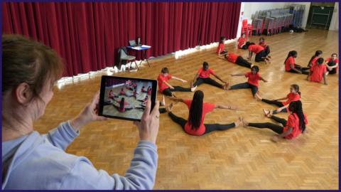 Secondary-school children in a school hall taking part in a dance lesson - they are sat on the wooden floor, facing each other in a circle. A teacher is holding a tablet and filming them.