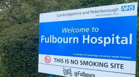 A blue and white sign saying Cambridgeshire and Peterborough NHS Foundation Trust, and Welcome to Fulbourn Hospital, standing on blue legs, against a dark green hedge, on a lighter green verge beside a road