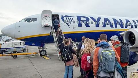 Passengers - mainly aged between 18-35 - boarding a Ryanair aircraft near the cockpit, via a mobile stairway on an airport tarmac under a cloudy sky. The aircraft is white with a blue and yellow stripe and the word ‘RYANAIR’ prominently displayed on the fuselage. People in casual clothing with backpacks and luggage are lined up, walking toward the plane’s front entrance.
