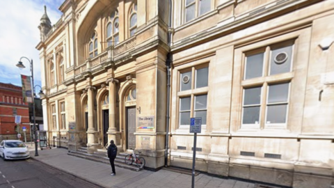 A large sandstone public building with columns and an ornamental balustrade over the entrance. 