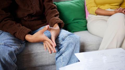 A stock image of a young person and a counsellor sat on a grey sofa. One person is wearing a brown jumper and blue jeans, and the other is wearing a yellow top and white trousers.
