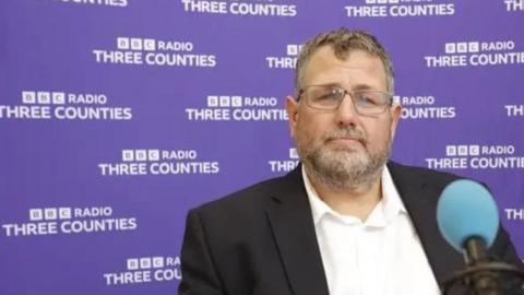 Peter Geary wearing a dark jacket and light-coloured shirt is seated in a radio studio in front of a microphone with a blue windscreen. The background is a purple branded backdrop repeating the text “BBC Radio Three Counties”. A second person is partially visible at the right edge of the image. The photograph is brightly lit and appears to have been taken during a radio interview.