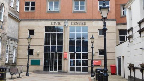 The front of a council building with large glass front windows with doors at the bottom. The building says "Civic Centre" above the windows. Old-fashioned street lamps are on either side of the path leading to the doors.