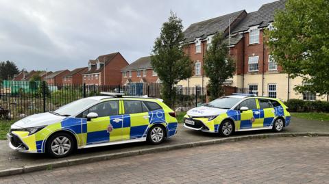 Two police cars with hi-vis yellow and blue decals, parked on the pavement on a residential estate. There is an allotment patch behind a black metal fence in the centre of the estate. In the background there is a row of flat buildings with a red brick façade on top. It is an overcast cloudy day. 