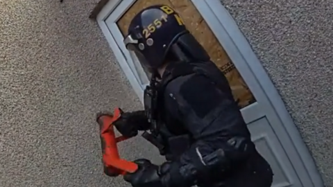 A police officer is using a red baton to smash through a boarded up white door. The house is pebble dashed. The officer is wearing a helmet and padded gear.