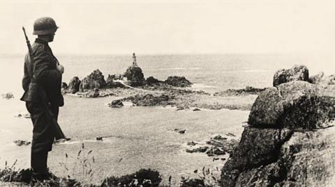 A German solider holding a rifle and wearing a tin helmet is standing on a cliff and looking out to sea