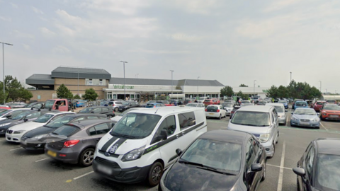 A google street view image of a Waitrose car park. There are cars and vans parked in the car park. In the background is a Waitrose building. There are trees and lampposts in the car park.