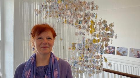 Gillian Taylor, who has red hair and is wearing purple, stands in front of her art installation. The daisies made from the letters hang on fishing wire from the ceiling of the stairwell.
