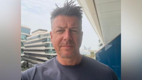 Luke Deal in a selfie-style image while he stands on a hotel balcony. He is slightly smiling at the camera. He has short dark hair with some grey. He wears a grey T-shirt.