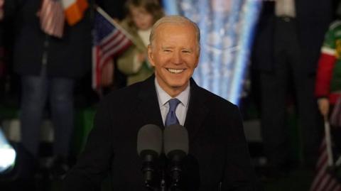 A man with white hair smiles. He is standing at a podium with an American eagle design on the front. He is wearing a suit. Behind him is a crowd, with a mixture of Irish and American flags. 