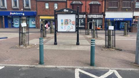 An empty town centre high street with several tree stumps encased in small black metal railings. There are shop fronts in the background.