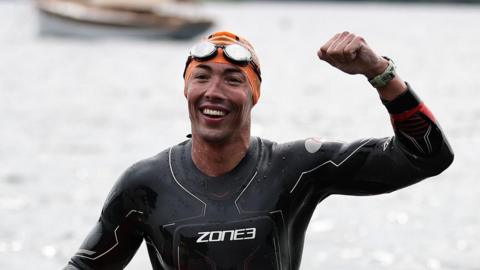 Adam Ball is coming out of the water, smiling broadly and raising his fist in victory. He is wearing an orange swim cap and a dark swimsuit.