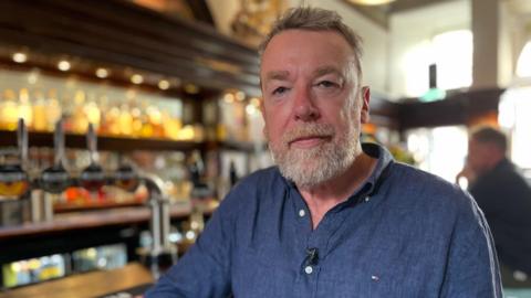 Alistair Brown looking straight at the camera. He has short grey hair and a white beard. He is wearing a blue shirt and is leaning on a bar with taps in the background.