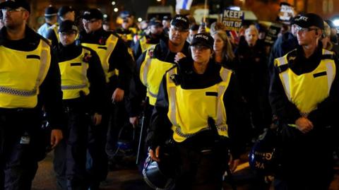 A line of police officers, all wearing hi-vis vests, walk in a line up a street. In the background a couple of protest banners can be seen.