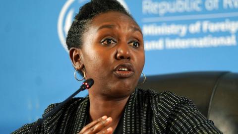 Rwanda government spokesperson Yolande Makole sits on a black chair before a microphone. She is wearing a white dotted black blazer.