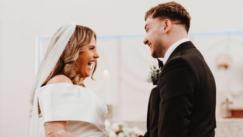 Caoimhe is smiling at Ollie. They are at the alter of their wedding. She is wearing a white veil and an off the shoulder wedding dress. He is wearing a dark coloured suit and has short dark hair. She has long blonde hair curled.