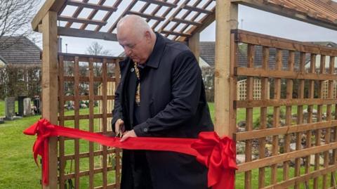 Lord Mayor of Stoke-on-Trent, Steve Watkins is stood under a wooden trellis with a red ribbon attached. He is holding a pair of scissors, and is cutting the ribbon.