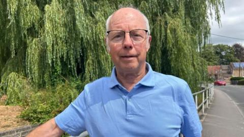 Councillor Paul McKernan, who has receding white hair and is wearing black-rimmed glasses and a light blue polo shirt, is pictured standing in front of a willow tree beside a road. His arm is resting on railings.