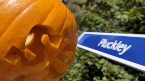 A carved pumpkin in front of a blue sign with the word Pluckley in white writing