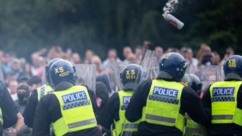 Riot police officers push back anti-migration protesters outside the Holiday Inn Express Hotel 