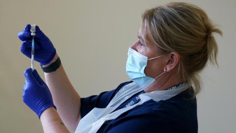A nurse dressed in blue and wearing a facemask, plastic apron and blue gloves is holding up a vaccine vial with the needle of a syringe in it. She has blonde, tied-back hair and has an NHS lanyard around her neck.
