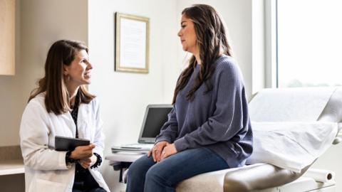 A Female doctor with mid length brown hair, wearing a white coat, holds an ipad  and discusses treatment with female patient who sits on a bed, and wears a blue jumper in an exam room