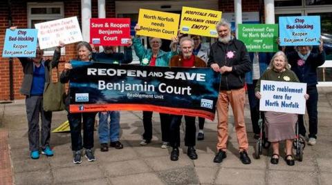 A brick building with a glass entrance with a group of protesters gathered outside holding banners reading "Save Benjamin Court".