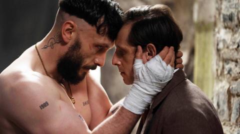 A man with a beard and bandages on his hands holds the face of a man wearing a suit.