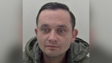 A police mugshot of a man with short black hair and green hoodie. 