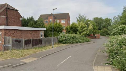 A road leads down to a brick wall and curves away from the camera. On the left is a building and a road sign saying Beck Gardens. Further along is a hedge and some small trees, with three-storey brick building behind them. On the right is a pavement area lines with bushes and trees.