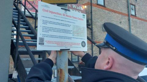 PCSO Daniel Grant putting up a civil injunction notice in St Neots, the sign is on a metal pole, with steps and a red brick building behind it. PCSO Grant is wearing a uniform and and hat and is standing away from the camera.