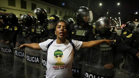A protester, wearing a white T-shirt with the slogan "police, you're poor, same as me", spreads her arms out at her side as she stands in front of a line of riot police holding shields. in Lima, Peru, on 28 September, 2025.