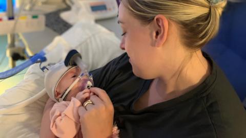 Maeva is in her mother's arms in hospital. Maeva is wearing a ventilator and has a white hat on and a pink top. Her mother, who has blonde hair, is looking lovingly at her baby.