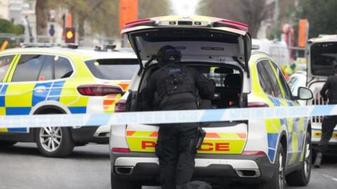 A firearms officer retrieving items from the boot of a police car. The firearms officer is dressed all in black and the boot of the police car is open. There is another police car to the left, with both vehicles and the officer standing behind a police cordon.