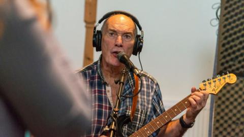 Dr Curran sings and plays the guitar in a studio. He has a serious expression and is looking away from the camera. He wears large black headphones and a muilt-coloured checked shirt. 