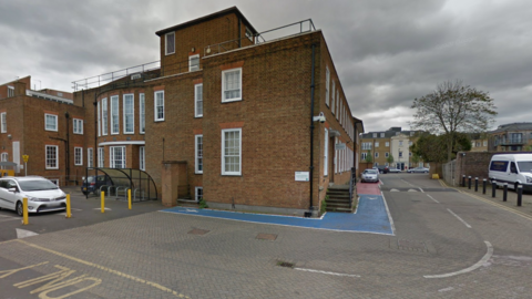 Shows the outside of a two-store red-brick building (the Epsom and Ewell Citizens Advice). Around the building are a few parked cars and a tree in the distance.